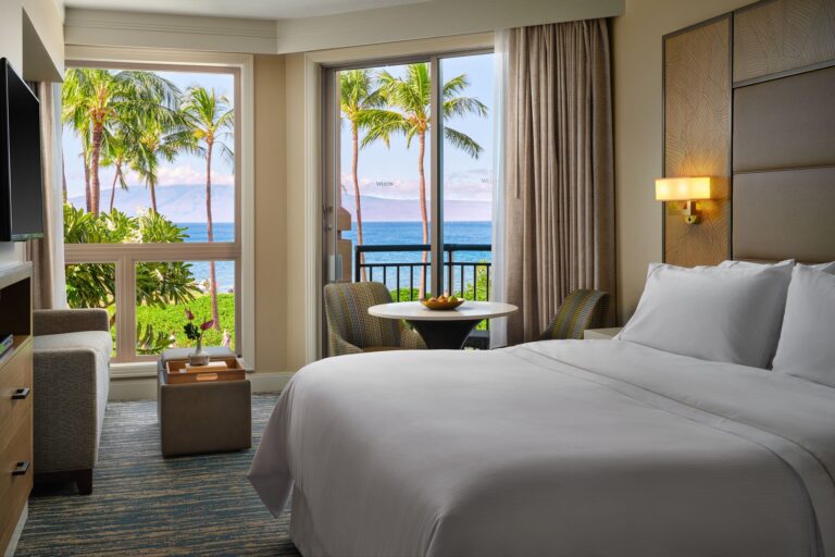Image of hotel bed with view of courtyard with palm trees and ocean.