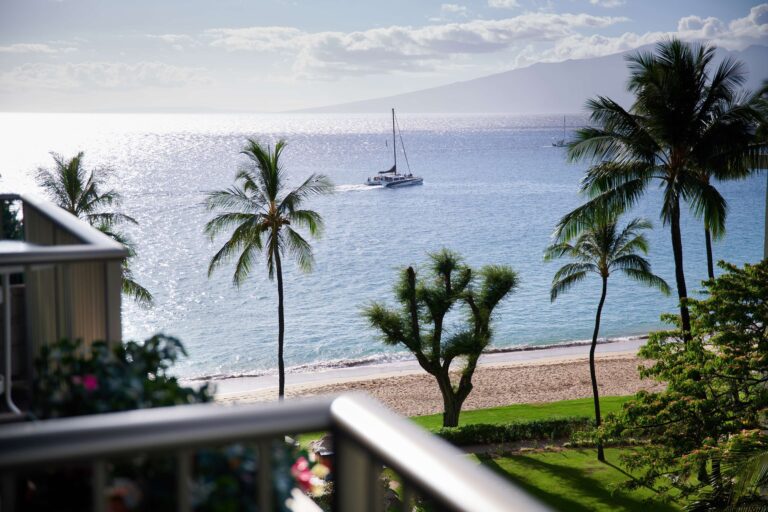 Image from island resort balcony with sail boat on ocean framed between palm trees.