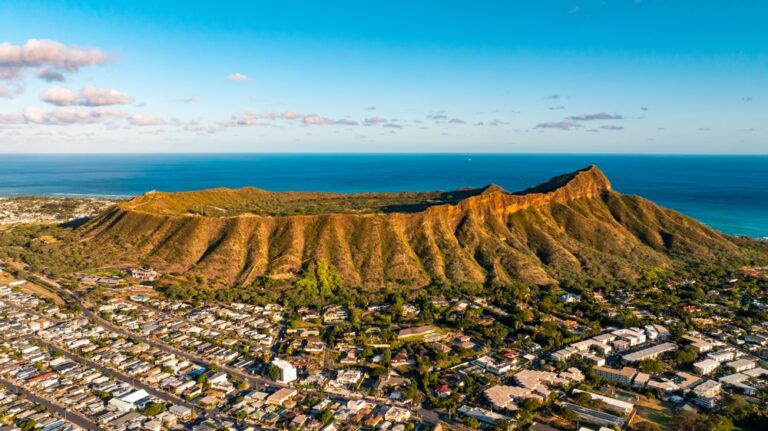 Rugged slopes of Diamond Head Crater are lit with bright sun. Cityscape of Honolulu around the rock. Aerial view.